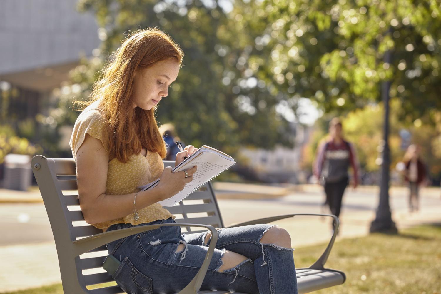 A <a href='http://sk.sundayhouse.net'>BETVLCTOR伟德登录</a> student reads on a bench along Campus Drive.